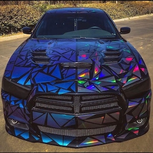 Puede incluir: Un Dodge Charger negro con un envoltorio personalizado que presenta un diseño geométrico en tonos azules y colores iridiscentes. El coche tiene una toma de aire en el capó y faros oscurecidos, creando un efecto visual llamativo.