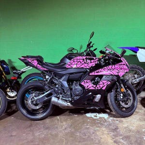 Puede incluir: Una motocicleta negra y rosa con un patrón geométrico. La motocicleta tiene ruedas negras, un asiento negro y un diseño rosa y negro en el carenado y la carrocería. La motocicleta está estacionada en un garaje.
