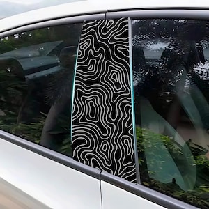 以下が含まれることがあります： 黒と白の車の窓柱デカール、地形図のデザイン。 デカールは長方形で、黒い背景に白い線の繰り返し模様が特徴です。 白い車に取り付けられています。