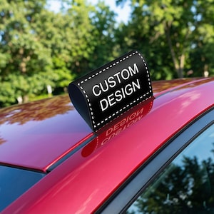 May include: A black, curved sticker with the words "CUSTOM DESIGN" in white, affixed to the red surface of a car. The sticker's edge is outlined with a dotted white line. The background features green foliage.