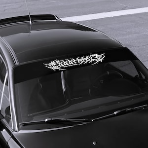 Op de afbeelding: Een zwarte auto met een witte afbeelding over de bovenkant van de voorruit. De afbeelding is een abstract ontwerp met scherpe, stekelige randen. De auto staat op een grijs oppervlak.