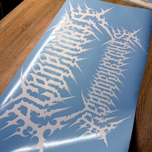 May include: A light blue vinyl sticker featuring a white, stylized design. The design has a spiky, organic shape with intricate details. The sticker is on a wooden surface, with other stickers visible in the background.
