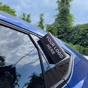 May include: A black sign with the text "YOUR LOGO HERE" is attached to the window of a blue car. The sky and trees are reflected in the car window.