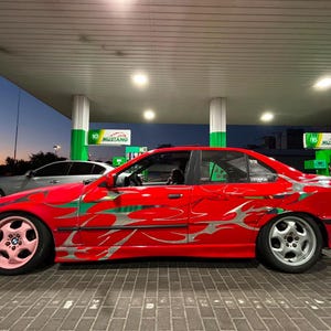 May include: A red saloon car featuring silver flame-style decals along the side panels. The front wheel has a pink five-spoke rim, while the rear wheel features a silver five-spoke rim. The vehicle is parked at a petrol station with green pillars.