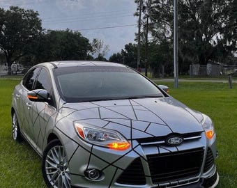 May include: Silver Ford Focus with a geometric black pattern across the hood and front panels. The car has a black front spoiler, tinted windows, and custom wheels. The text "FORD PERFORMANCE" is visible on the windshield.