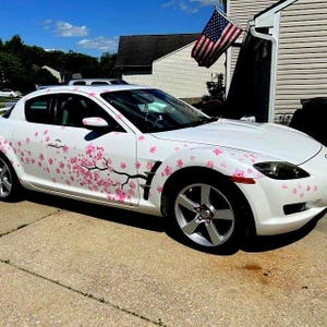 Könnte beinhalten: Weißes Sportauto mit rosa Kirschblüten-Aufklebern. Das Auto hat silberne Felgen und steht auf einer Betonauffahrt. Im Hintergrund ist eine amerikanische Flagge zu sehen.