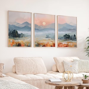 Puede incluir: Tres pinturas de paisajes enmarcadas con un tema de puesta de sol. La obra de arte presenta montañas, árboles y campos en tonos azules, naranjas y amarillos. Los marcos son de color madera clara. Las pinturas están sobre un sofá seccional beige.