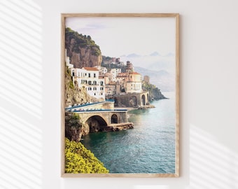 May include: A framed photograph of a coastal town in Italy. The town is built on a cliffside overlooking the turquoise blue water. The photo features a stone arch bridge leading to the town.