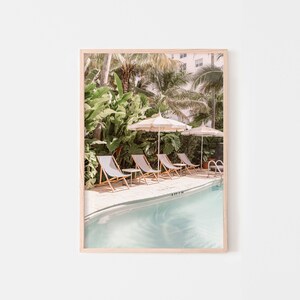 May include: A framed print featuring a serene pool scene. The image showcases a row of lounge chairs, striped umbrellas, and lush greenery. The pool's water has a light blue hue, creating a relaxing atmosphere. The print is in a light wooden frame.