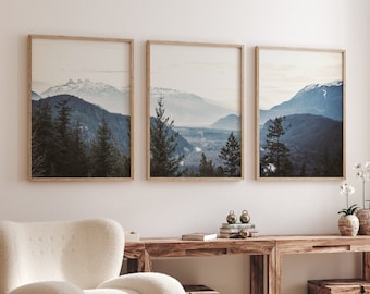 May include: Three framed landscape photographs of a mountain range with snow-capped peaks and a valley with trees. The photographs are hanging on a white wall above a wooden table with a white armchair and a round wooden coffee table.