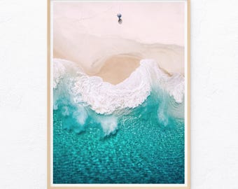 May include: A framed aerial photograph of a sandy beach meeting turquoise ocean waves. A single small blue umbrella sits on the white sand, with faint footprints leading toward it, captured from a high vertical perspective in a light wood frame.
