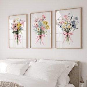 May include: Three framed watercolour prints of floral bouquets. Each bouquet features a variety of colourful flowers, including pink, yellow, and blue blooms, tied with a pink ribbon. The frames are light wood, displayed above a bed with white bedding.