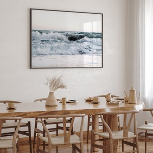 Op de afbeelding: Een houten eettafel met zes stoelen, een vaas met gedroogde bloemen en een ingelijst foto van oceaan golven die breken op een strand.