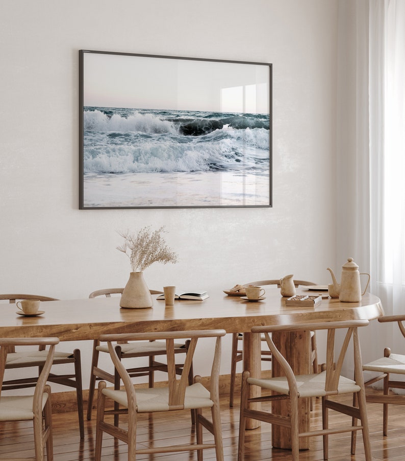 Op de afbeelding: Een houten eettafel met zes stoelen, een vaas met gedroogde bloemen en een ingelijst foto van oceaan golven die breken op een strand.
