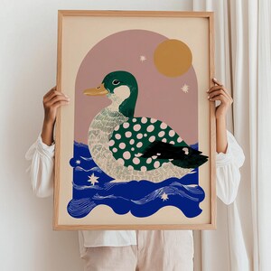 May include: A framed art print depicting a stylized duck with green and pink polka dots, on blue water. The background features a pink arch, a yellow sun, and stars. The frame is made of light-coloured wood.