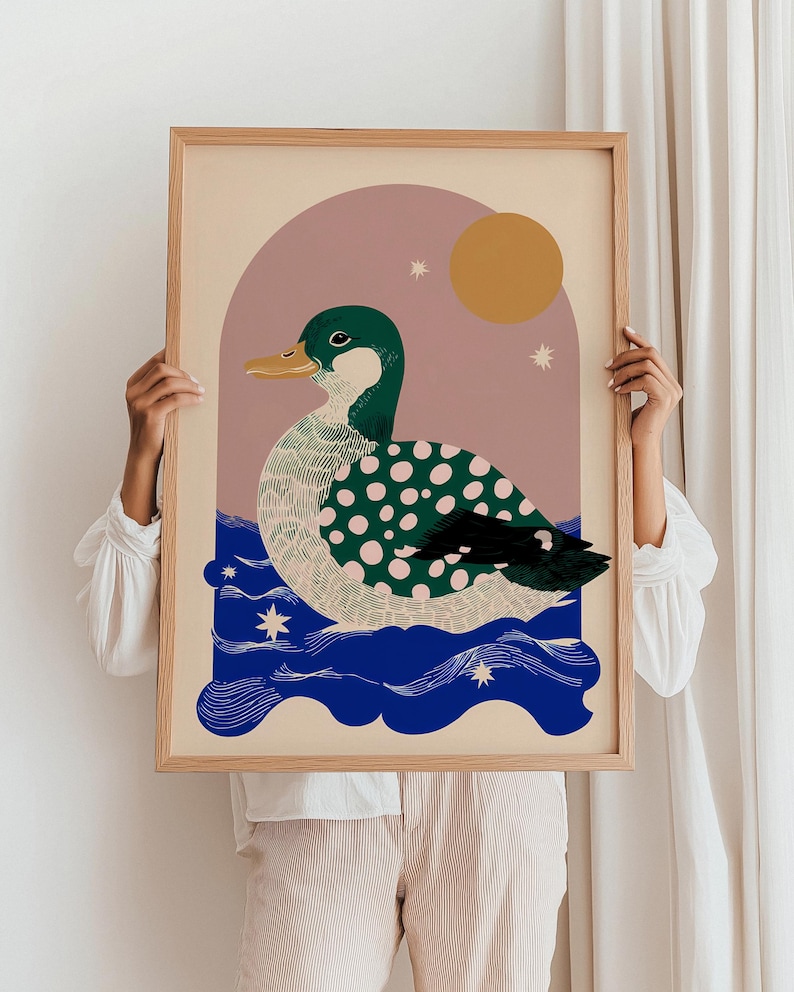May include: A framed art print depicting a stylized duck with green and pink polka dots, on blue water. The background features a pink arch, a yellow sun, and stars. The frame is made of light-coloured wood.
