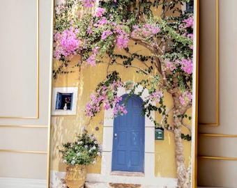 Peut inclure: Un bâtiment jaune avec une porte bleue et une vigne fleurie rose qui pousse dessus. La porte a une arche décorative et une petite fenêtre au-dessus. Le bâtiment est entouré d'une végétation luxuriante.