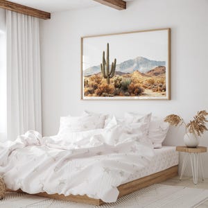 May include: A bedroom scene featuring a framed desert landscape art piece above a bed with white bedding. The bedding has a star pattern. A small wooden side table with a vase of dried plants sits beside the bed. The room has white walls and a white rug.
