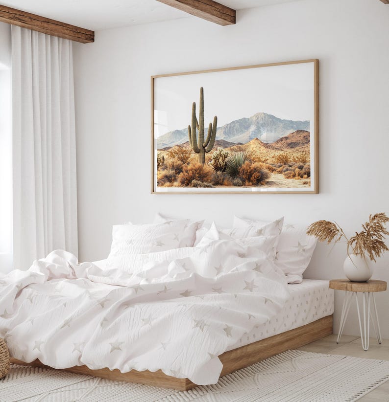 May include: A bedroom scene featuring a framed desert landscape art piece above a bed with white bedding. The bedding has a star pattern. A small wooden side table with a vase of dried plants sits beside the bed. The room has white walls and a white rug.