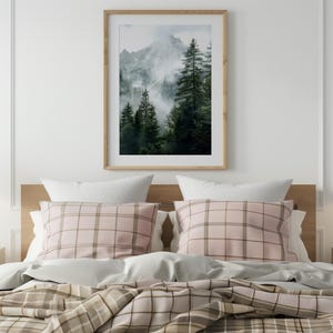 May include: A bedroom with a bed with a pink and brown plaid duvet cover and a framed photograph of a mountain scene with trees and fog.
