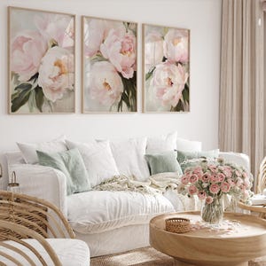 May include: Three framed floral art prints featuring soft pink peonies with green foliage. The artwork hangs above a white sofa with decorative pillows and a wooden coffee table. A vase of pink flowers sits on the table.