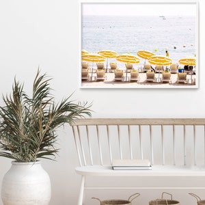 Puede incluir: Una impresi&oacute;n enmarcada de una escena de playa con sombrillas amarillas y tumbonas. El oc&eacute;ano est&aacute; al fondo. La impresi&oacute;n se exhibe en una pared blanca sobre un banco blanco con cestas tejidas y un jarr&oacute;n blanco grande con vegetaci&oacute;n.
