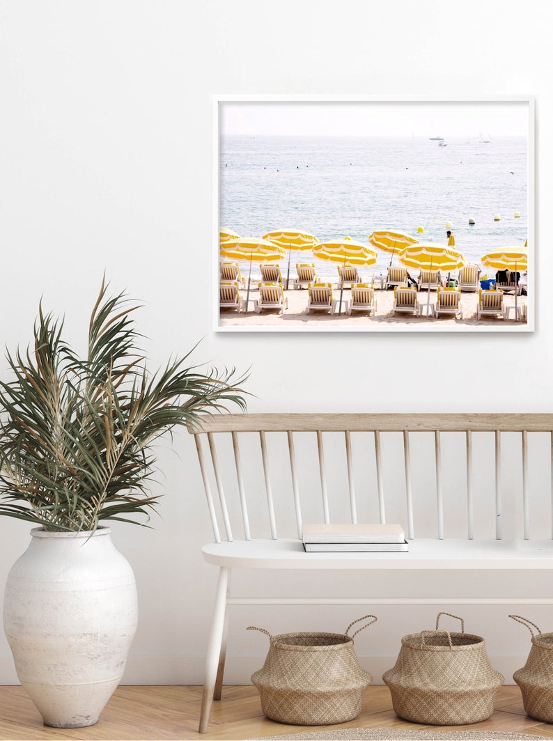 Puede incluir: Una impresi&oacute;n enmarcada de una escena de playa con sombrillas amarillas y tumbonas. El oc&eacute;ano est&aacute; al fondo. La impresi&oacute;n se exhibe en una pared blanca sobre un banco blanco con cestas tejidas y un jarr&oacute;n blanco grande con vegetaci&oacute;n.