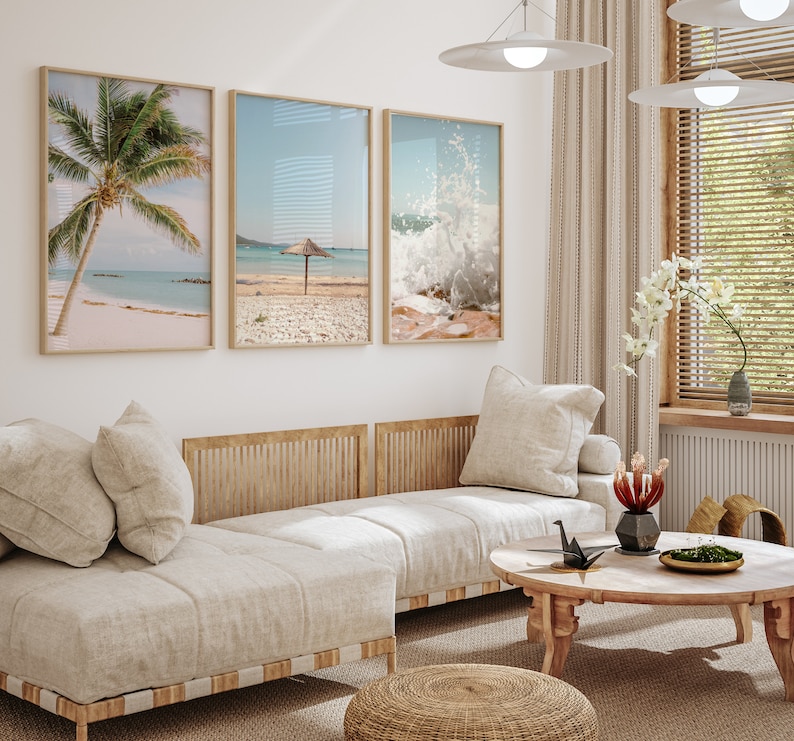 May include: Three framed beach-themed prints with a tropical palm tree, a beach with an umbrella, and a wave crashing on the shore. The prints are hanging on a white wall above a light beige couch with a wooden frame.