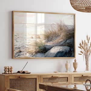 Könnte beinhalten: Gerahmte Landschaftsfotografie einer Strandlandschaft. Das Bild zeigt Sanddünen mit hohem, trockenem Gras, einen Sandstrand und einen Blick auf das Meer unter einem hellen Himmel. Der Rahmen besteht aus hellem Holz.