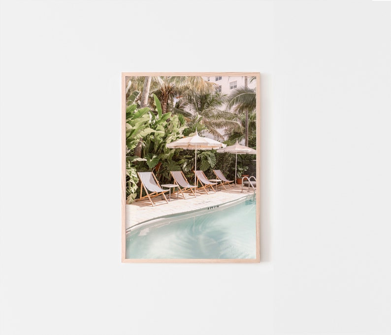 May include: A framed print featuring a serene pool scene. The image showcases a row of lounge chairs, striped umbrellas, and lush greenery. The pool's water has a light blue hue, creating a relaxing atmosphere. The print is in a light wooden frame.