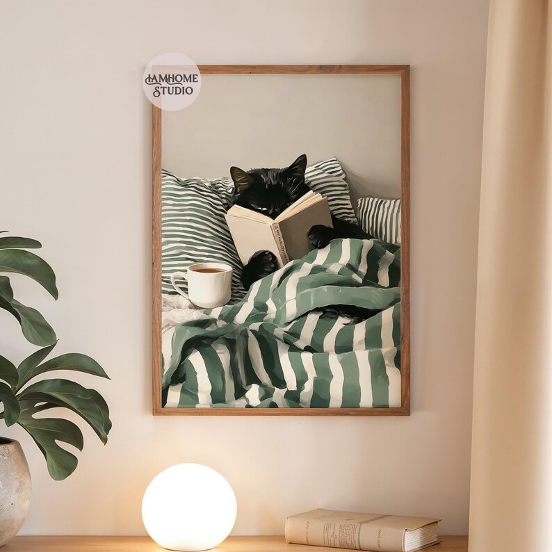 May include: A framed print depicts a black cat reading a book in bed, nestled under a green and white striped blanket. A white mug of tea sits nearby. The print has a wooden frame and is mounted on a neutral wall.