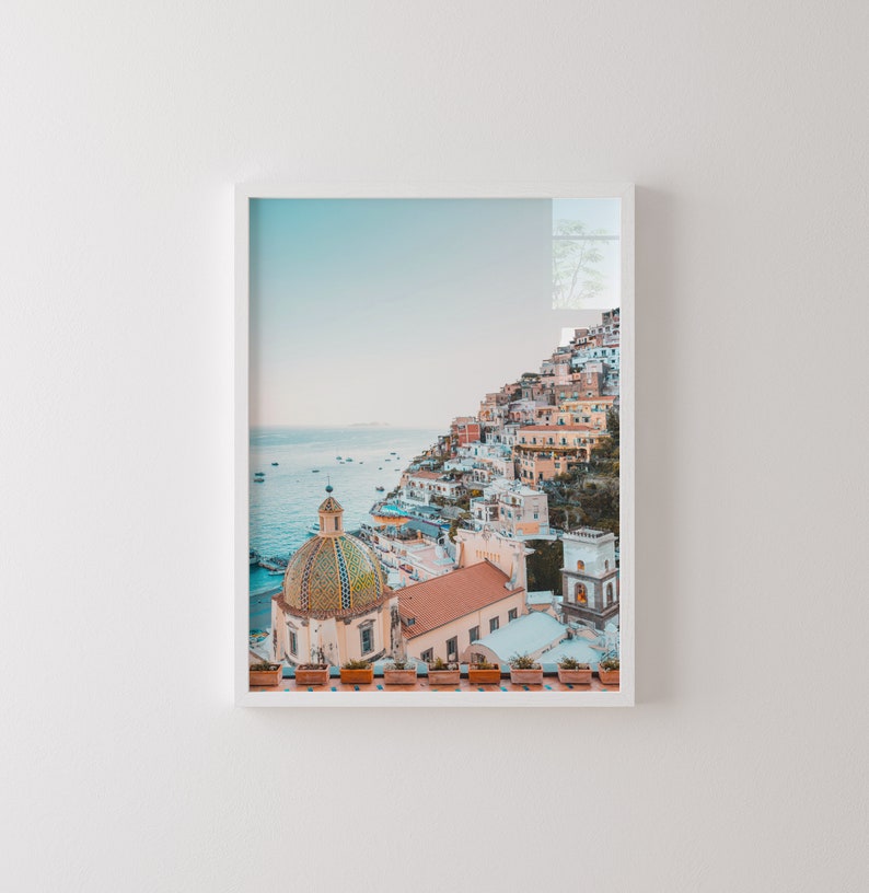 May include: A colorful cityscape with a white domed church building on a cliff overlooking the ocean. The buildings are painted in shades of pink, yellow, and orange. The ocean is a light blue color and there are boats in the water.