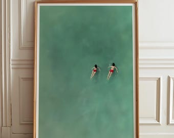 May include: A framed art print featuring two figures swimming in turquoise water. The figures are wearing red swimwear. The artwork is displayed in a wooden frame, set against a white wall and a parquet floor.