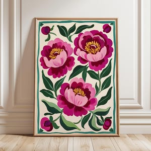 May include: A framed print featuring a floral design with three pink and magenta peonies on a cream background. The peonies have yellow centers and are surrounded by green leaves. The frame is a light wood color with a teal border.