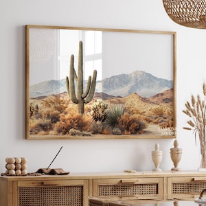 May include: A framed print featuring a desert landscape with a large saguaro cactus in the center. The artwork showcases a mountain range in the background, with various desert plants and shrubs in the foreground. The color palette includes warm tones of brown, beige, and green.
