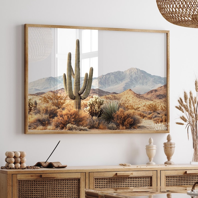 May include: A framed print featuring a desert landscape with a large saguaro cactus in the center. The artwork showcases a mountain range in the background, with various desert plants and shrubs in the foreground. The color palette includes warm tones of brown, beige, and green.