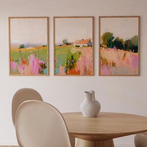 May include: A set of three framed landscape paintings featuring fields with pink and green brushstrokes, a white house with a red roof, and trees. The artwork is displayed in light wood frames, accompanied by a white ceramic vase on a wooden table.