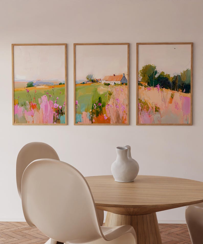 May include: A set of three framed landscape paintings featuring fields with pink and green brushstrokes, a white house with a red roof, and trees. The artwork is displayed in light wood frames, accompanied by a white ceramic vase on a wooden table.