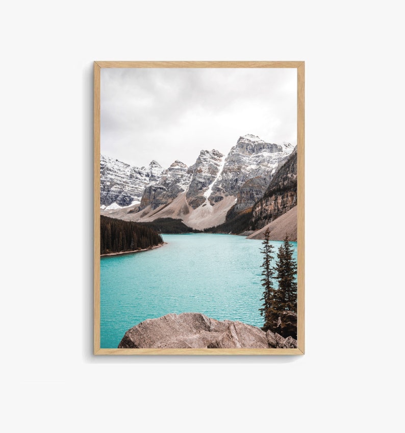 May include: A scenic photograph of a turquoise lake surrounded by snow-capped mountains. The lake is calm and reflects the sky, and there are a few trees in the foreground.