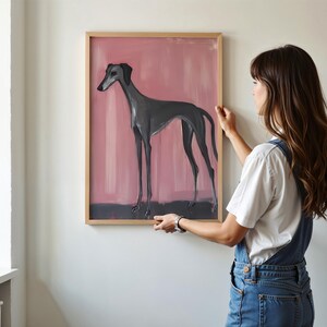 Puede incluir: Obra de arte enmarcada con una ilustraci&oacute;n de un galgo. El perro est&aacute; representado en gris sobre un fondo rosa. La obra est&aacute; encerrada en un marco de madera marr&oacute;n claro, proporcionando una est&eacute;tica cl&aacute;sica.
