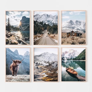 May include: A set of six framed wall art prints featuring mountain landscapes. Images include a sunlit lake, a road through a forest, a rustic cabin, a highland cow, a snowy valley with a winding stream, and a wooden rowboat on a calm lake.
