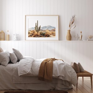 May include: A bedroom scene featuring a framed desert landscape print above a bed with white bedding and a brown throw blanket. Decorative items on a shelf include a woven vase, glass bottles, and a coral sculpture. The room has a neutral color palette.