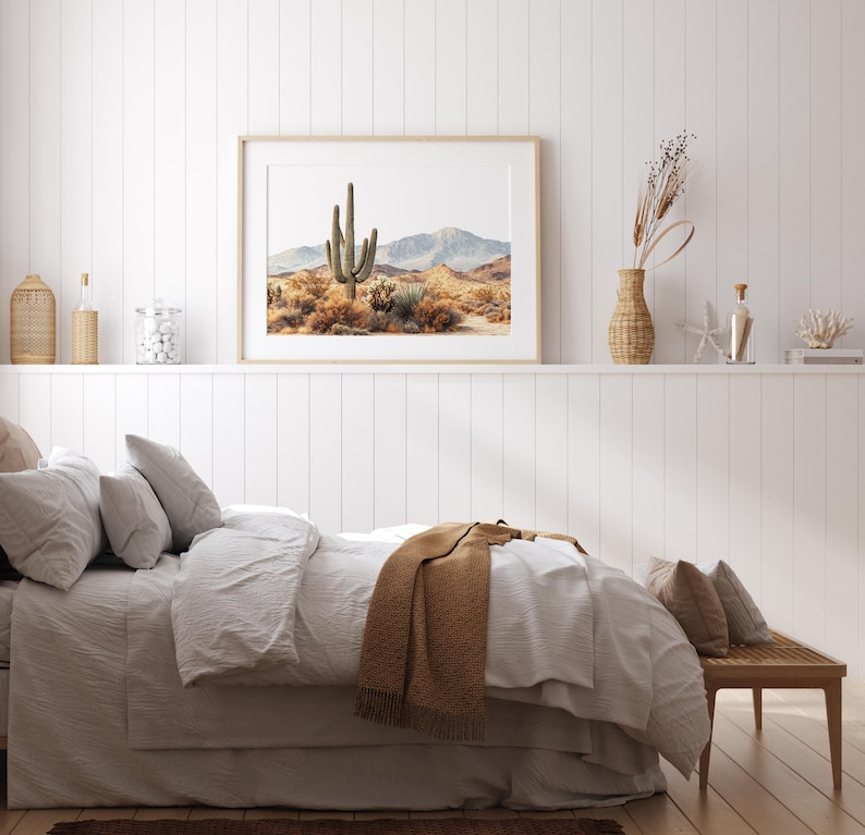 May include: A bedroom scene featuring a framed desert landscape print above a bed with white bedding and a brown throw blanket. Decorative items on a shelf include a woven vase, glass bottles, and a coral sculpture. The room has a neutral color palette.