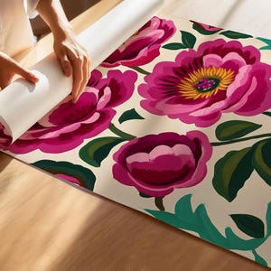 Puede incluir: Un rollo de papel tapiz con estampado floral rosa y verde sobre fondo blanco. El papel tapiz est&aacute; siendo desenrollado por una mano.
