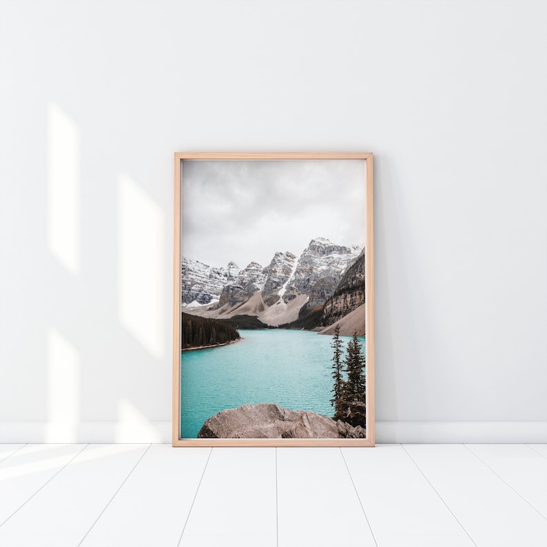 May include: A framed photograph of a mountain lake with snow-capped peaks in the background. The lake is a vibrant turquoise blue and the mountains are a soft white and gray. The photo is framed in a simple wooden frame.