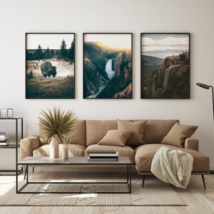 May include: Three framed landscape photographs hanging on a white wall above a brown sectional sofa. The photos feature a bison in a field, a waterfall in a canyon, and a mountain vista.