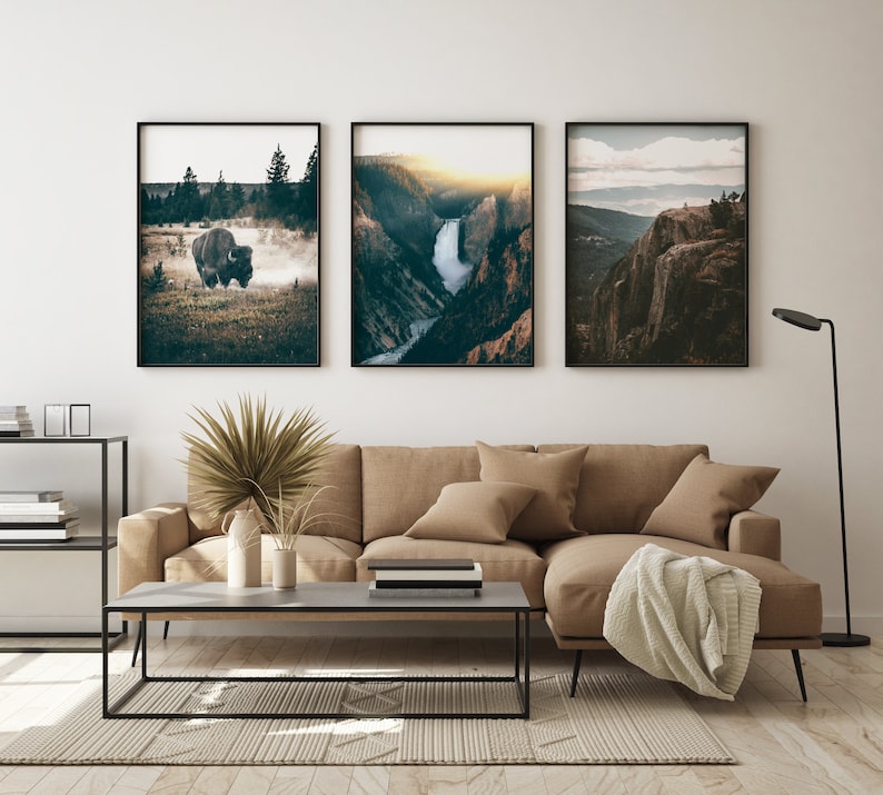 May include: Three framed landscape photographs hanging on a white wall above a brown sectional sofa. The photos feature a bison in a field, a waterfall in a canyon, and a mountain vista.