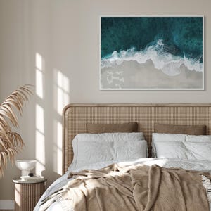 May include: A framed aerial photograph of a beach scene. The image captures the turquoise ocean waves crashing onto a sandy shore. The photograph is displayed above a bed with a woven headboard and neutral-toned bedding.