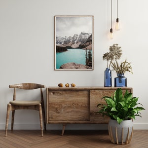 May include: A wooden cabinet with a sliding door, a wooden chair, a potted plant, and a framed photograph of a mountain lake. The cabinet is brown and has a natural wood finish. The chair is also brown and has a natural wood finish. The plant is green and has large leaves. The photograph is of a mountain lake with snow-capped mountains in the background.