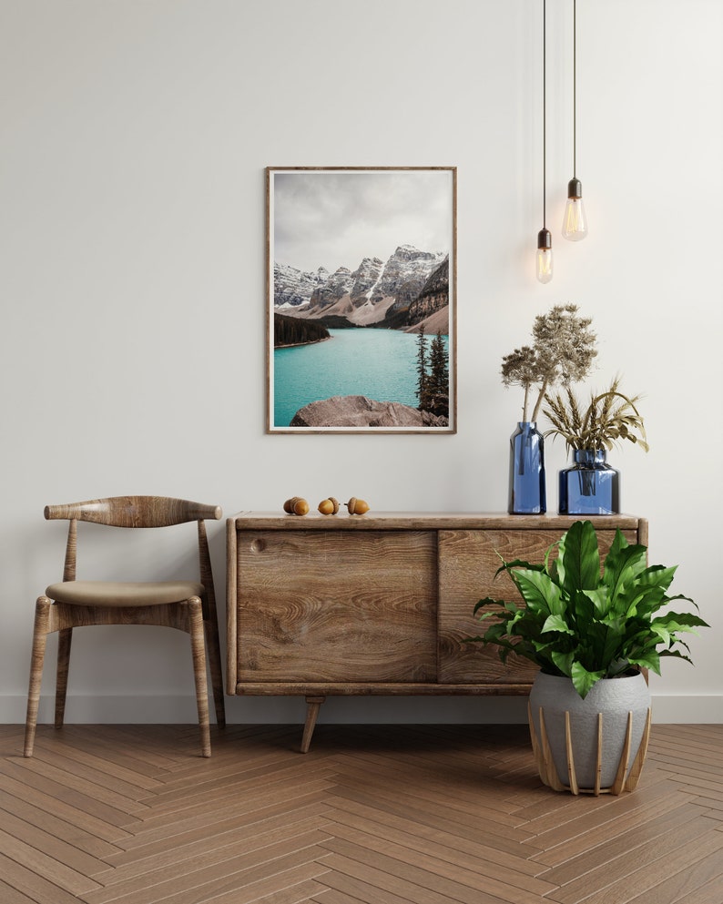 May include: A wooden cabinet with a sliding door, a wooden chair, a potted plant, and a framed photograph of a mountain lake. The cabinet is brown and has a natural wood finish. The chair is also brown and has a natural wood finish. The plant is green and has large leaves. The photograph is of a mountain lake with snow-capped mountains in the background.